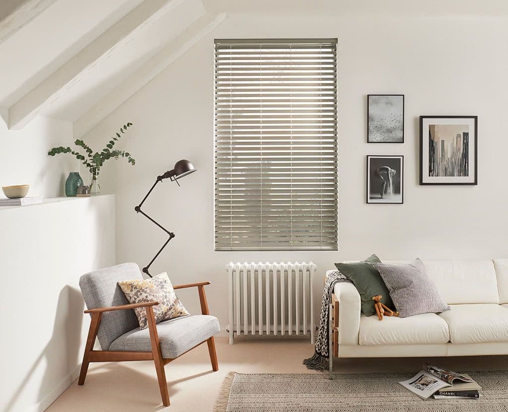 Horizontal aluminum Venetian blinds covering the central window, mostly closed and softly filtering daylight; cozy living room with a sofa, armchair, floor lamp, radiator, framed art, and neutral decor.