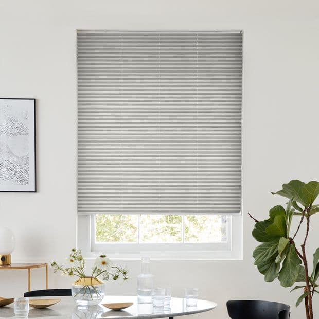 Light-gray pleated blind, lowered to just above the sill with horizontal folds, gently filtering daylight; covering a window in a bright dining area with round table, glassware, and potted plant.