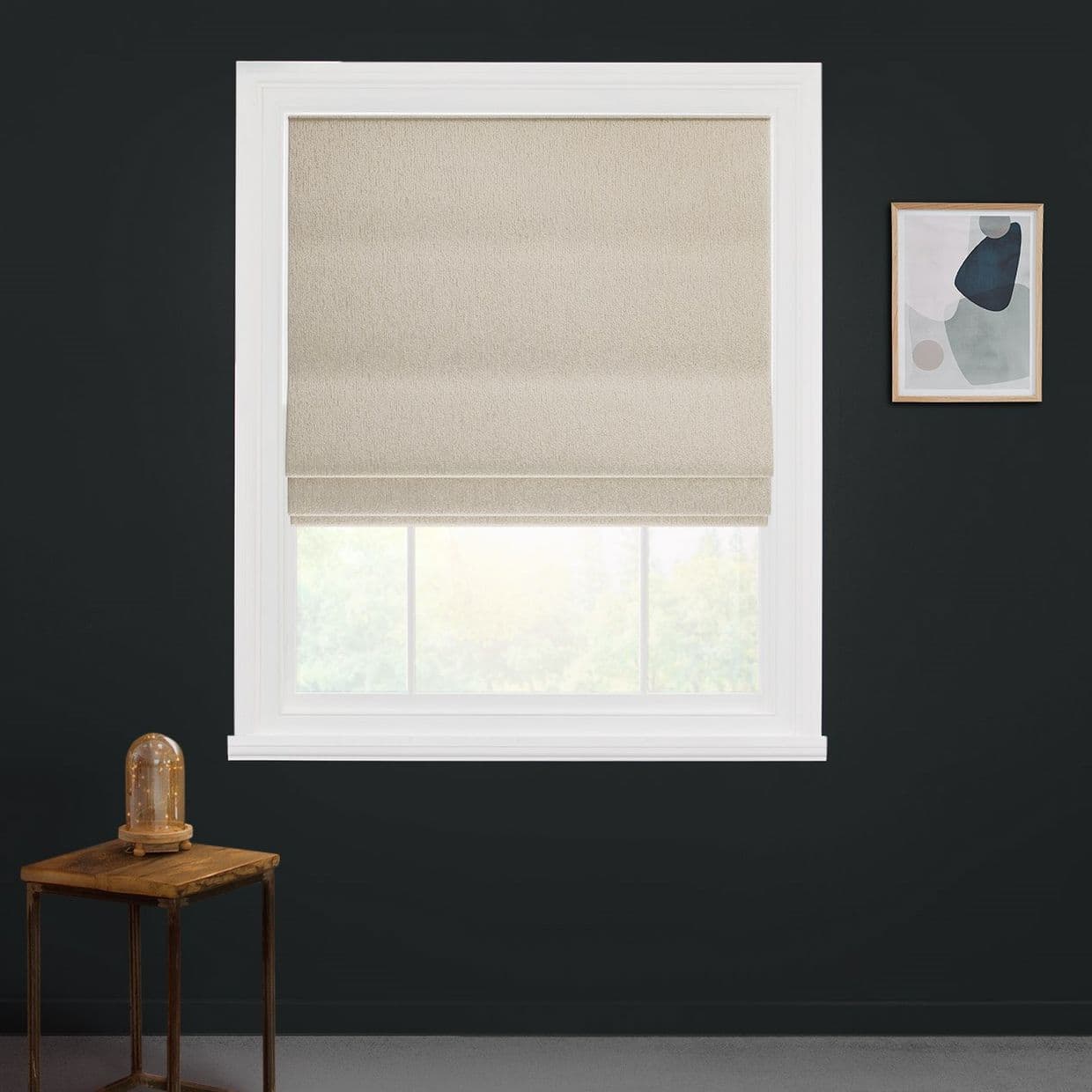 Beige fabric Roman blind centered in white-trimmed window, lowered midway and softly filtering daylight; set against a dark charcoal wall with a small wooden side table and abstract framed art.