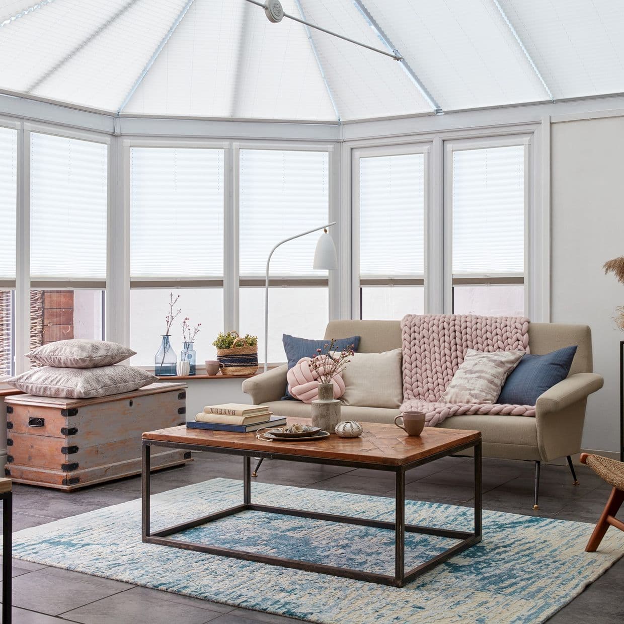 White pleated cellular blinds covering conservatory roof and tall bay windows, fully lowered and softly filtering daylight; a sunlit sitting room with beige sofa, chunky pink throw, and wooden coffee table.