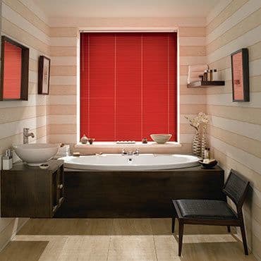 Red horizontal blinds centered over a window, fully closed and dimming light; above a white oval bathtub in a warmly lit striped-wood bathroom with sink, mirror, shelf and chair.