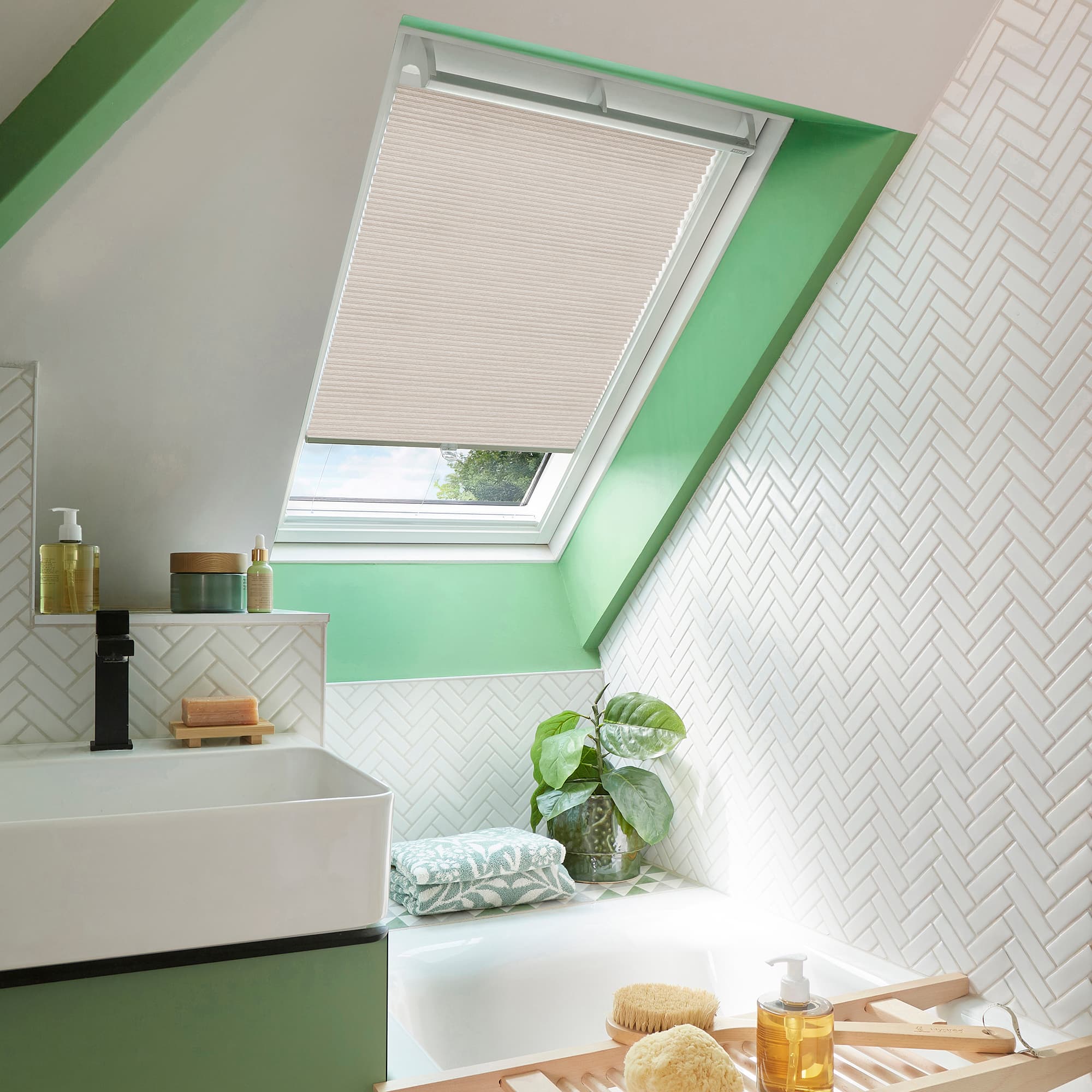 A beige pleated cellular blind covers a skylight, lowered halfway and filtering daylight; set in a mintâ€‘green framed, slopedâ€‘ceiling bathroom with white herringbone tiles, sink, bathtub, plant and bath tray.