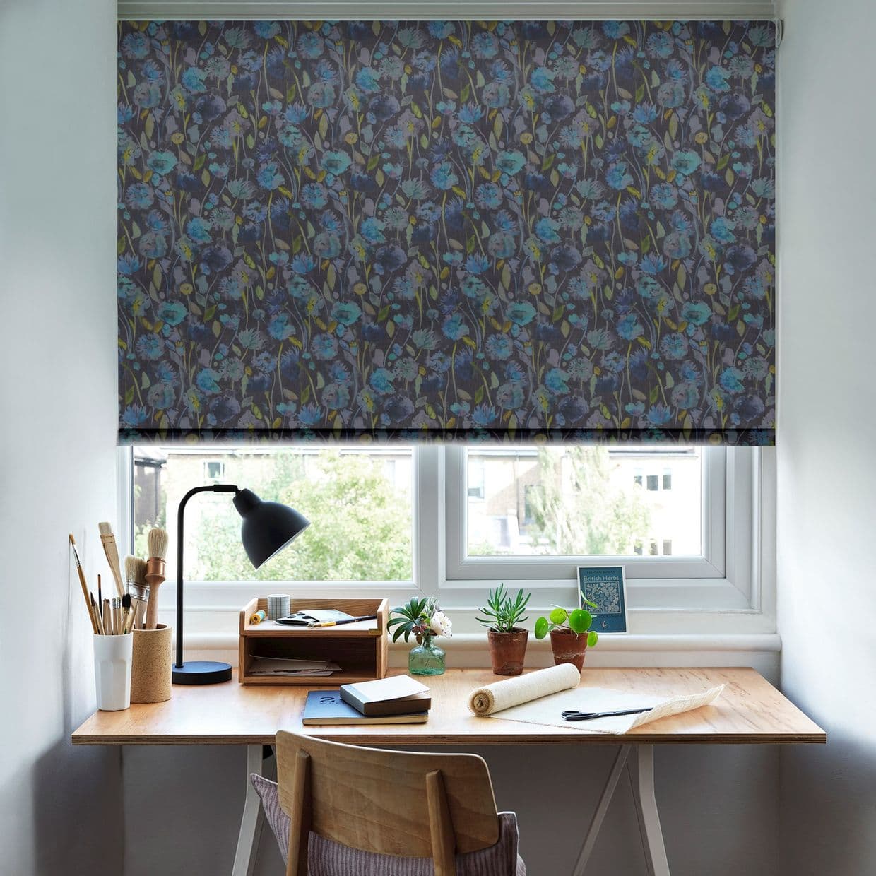 A dark floral roller blind, lowered across the window with blue flowers, filtering daylight; above a wooden desk with lamp, paintbrushes, potted plants and notebooks, light visible through a gap.