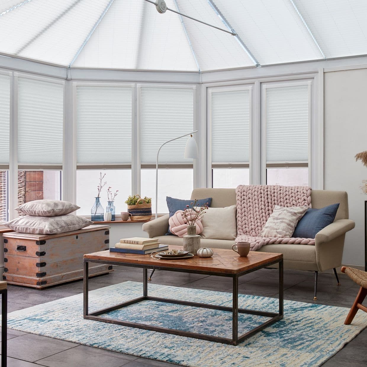 White pleated cellular blinds cover conservatory windows and skylight, mostly lowered, filtering daylight; a cozy living room with beige sofa, pink chunky throw, wooden coffee table and blue rug.