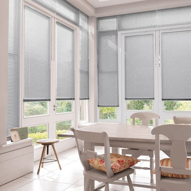 Grey horizontal slat venetian blinds covering floor-to-ceiling French windows and doors, partially lowered and filtering soft daylight; dining area with white wooden table, chairs and floral cushions, garden view beyond.