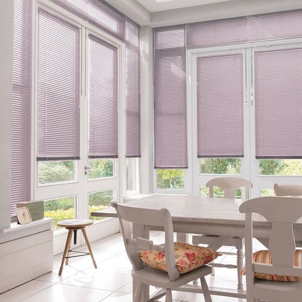 Pleated mauve cellular blinds covering tall white-framed French doors and windows, lowered to mid-height and filtering bright daylight into a sunlit dining nook with white table, chairs, and garden view.