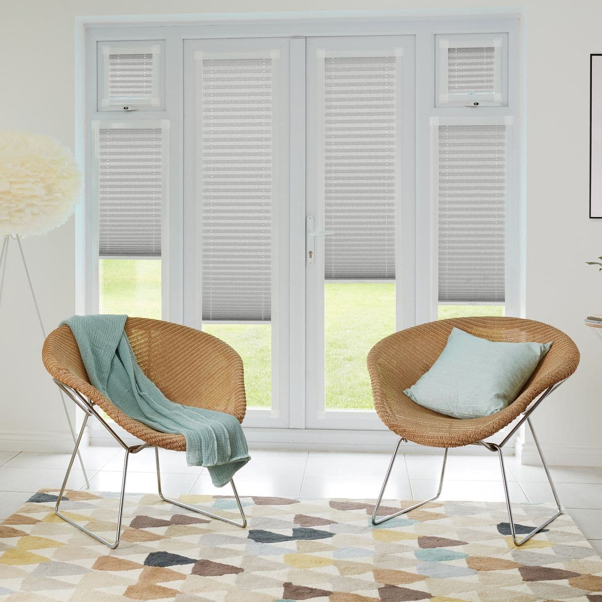 Grey honeycomb (cellular) pleated blinds cover tall French doors and sidelights, lowered mid-height and softly filtering daylight; bright sitting area with two wicker chairs, pastel cushions, patterned rug, lawn view.