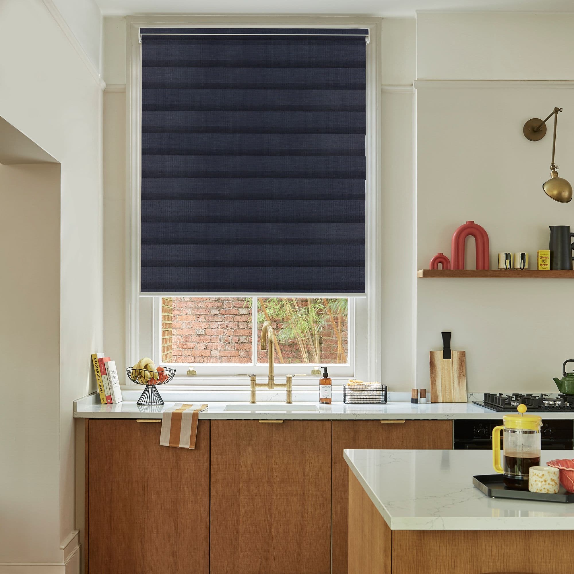 A navy dual-layer (zebra) roller blind lowered to mid-window, its horizontal bands filtering light over a kitchen sink with brass faucet, white countertop, wooden cabinets and open shelving.