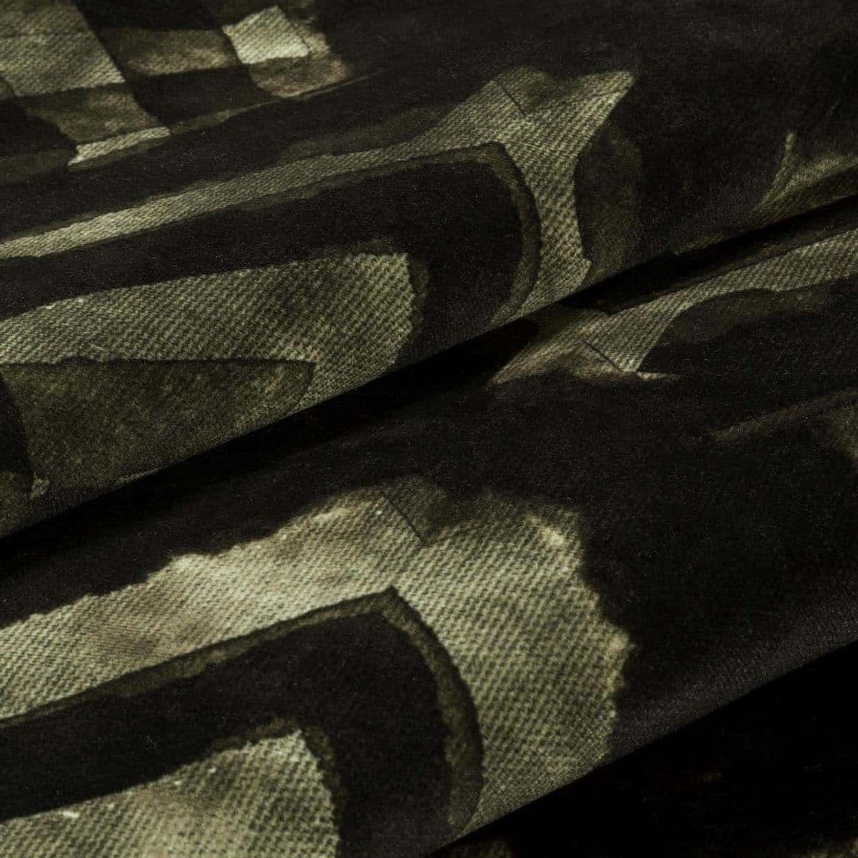 Velvet-like patterned fabric with dark olive and black abstract geometric print; folded into overlapping layers, showing soft nap and woven texture in dim, close-up studio lighting.
