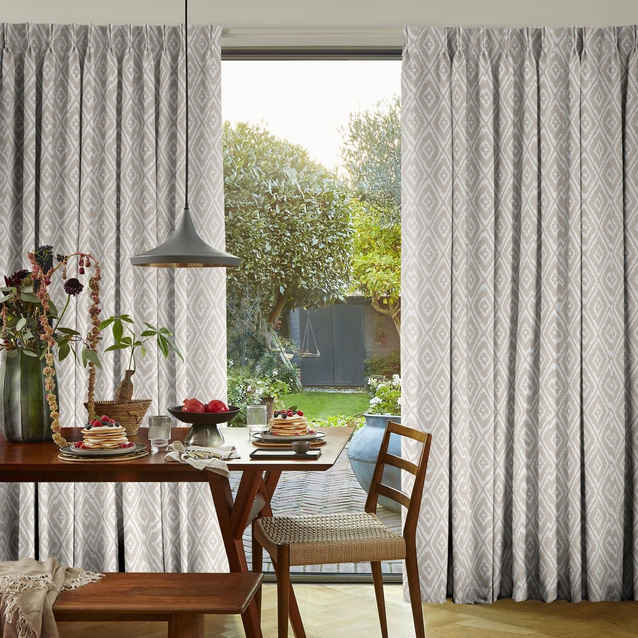 Pleated, full-length beige curtains with a white diamond pattern, parted to reveal a glass door; admitting bright daylight; dining table set for breakfast with pendant lamp, chairs, and garden beyond.