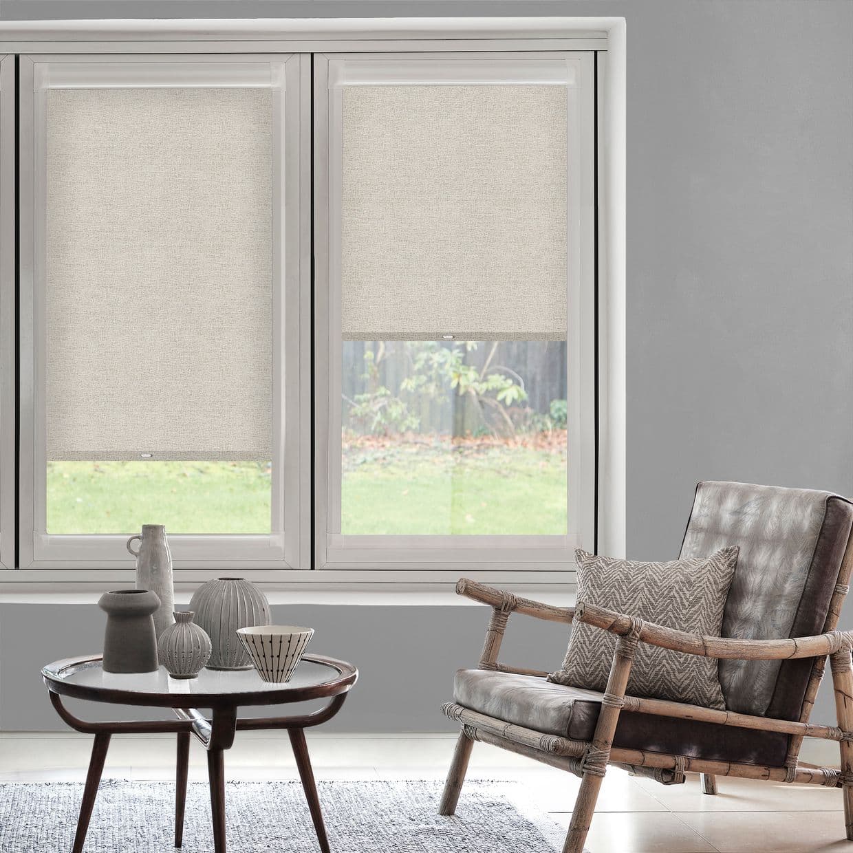 Beige fabric roller blinds cover twin windowsâ€”left fully lowered, right partially raisedâ€”filtering soft daylight. Modern gray living room with rattan armchair, patterned cushion, round coffee table and grassy backyard.