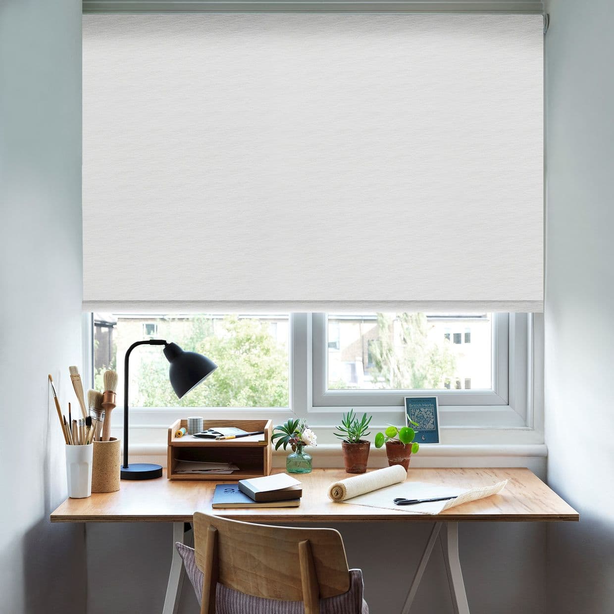 White fabric roller blind, fully lowered from a slim cassette, lightly filtering daylight. Small home workspace: wooden desk with lamp, brushes, notebooks and potted plants beneath the window, daytime light.