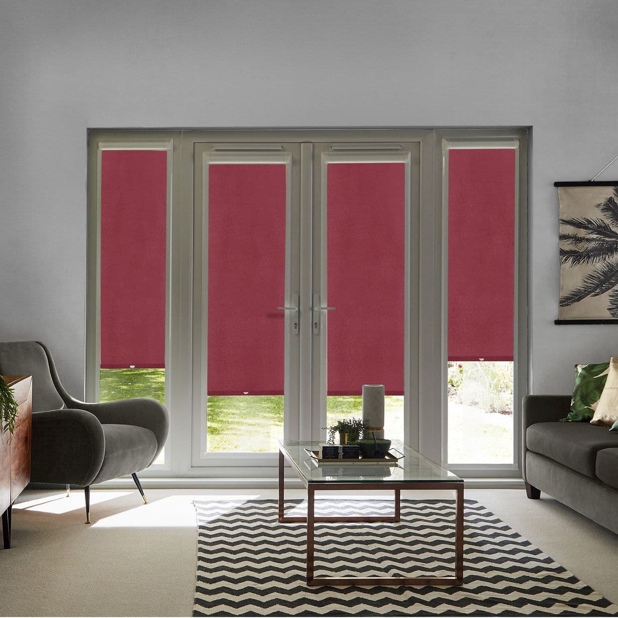 Red roller blinds cover four-panel French doors and sidelights, fully lowered and dimming light; living room with gray sofas, glass coffee table, chevron rug, garden visible below.