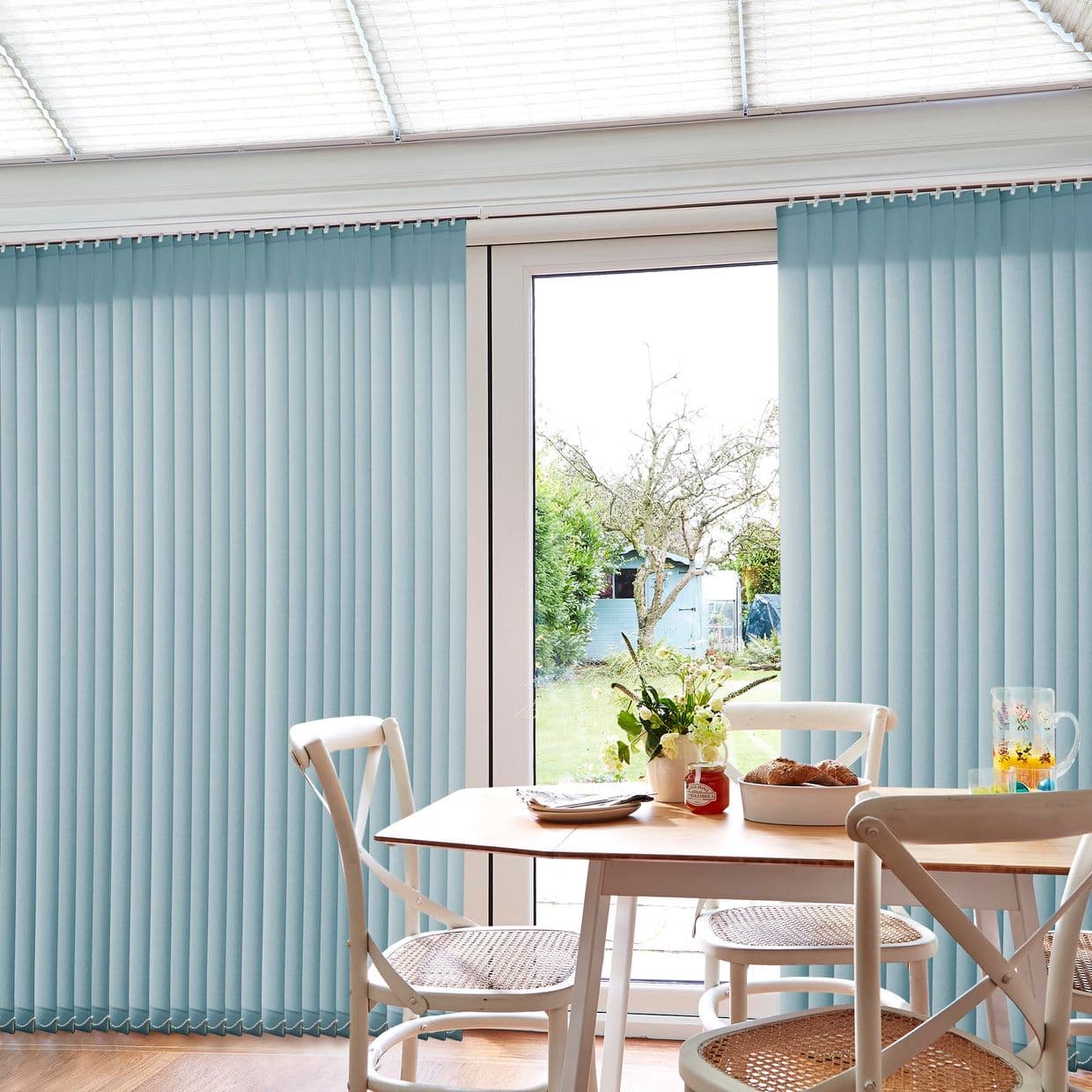 Light-blue vertical blinds on sliding glass doors, mostly closed with a central gap; filtering soft daylight, partly parted to reveal a backyard. Bright dining nook with wooden table and chairs.