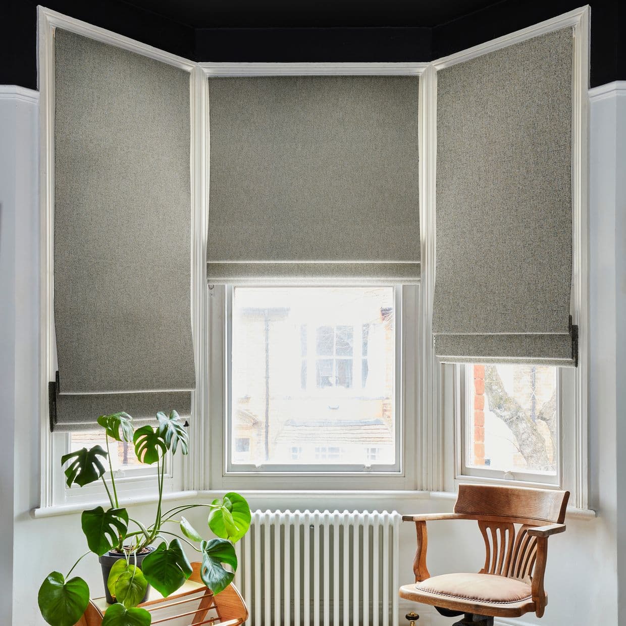 Grey fabric Roman blinds covering a three-panel bay window, partially lowered and filtering bright daylight into a white room with a radiator below, a wooden armchair, and a potted Monstera.