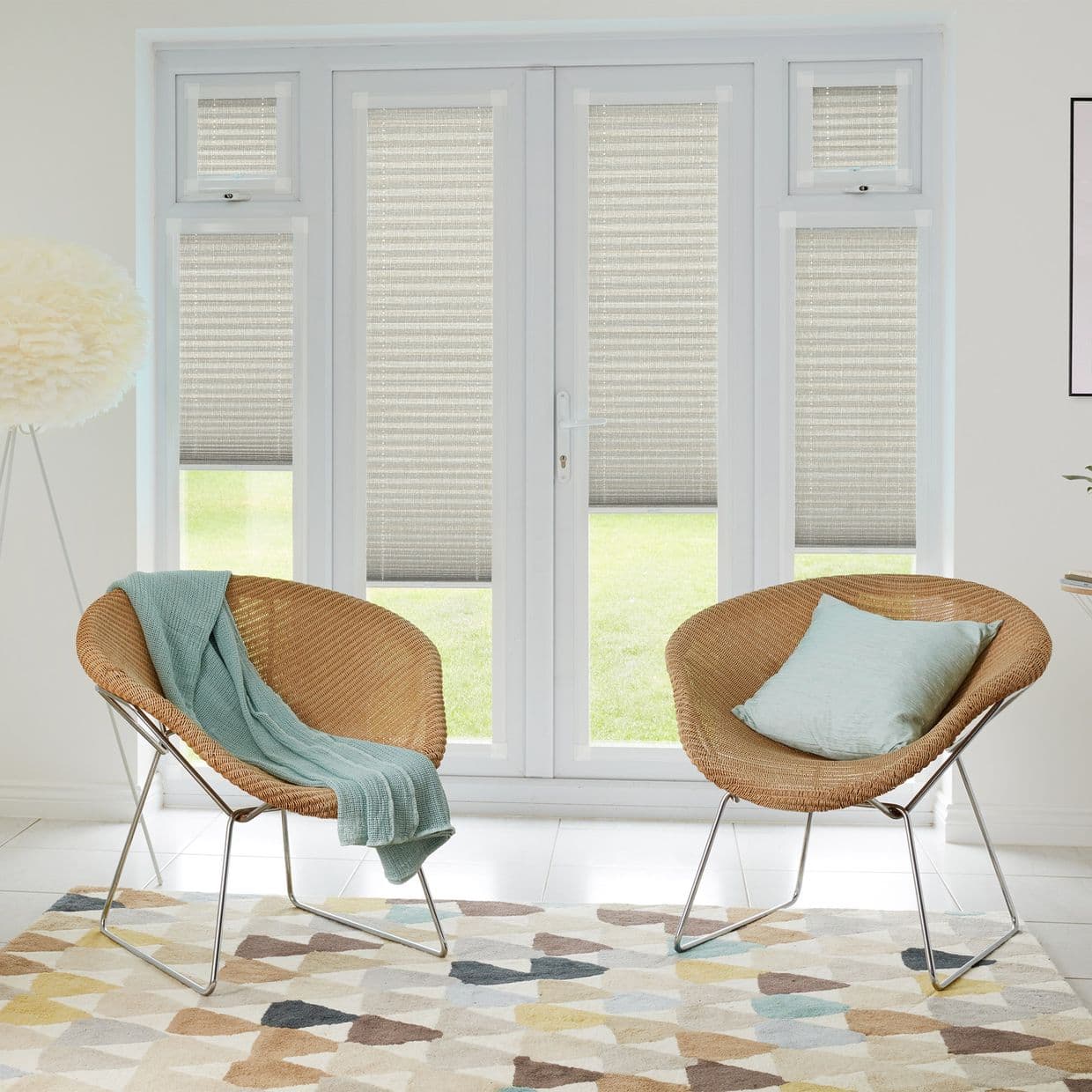 Beige pleated cellular blinds on French doors and sidelights, lowered mid-height and filtering daylight; bright living room with two wicker chairs, pale-blue throw and cushion, geometric rug, lawn visible outside.