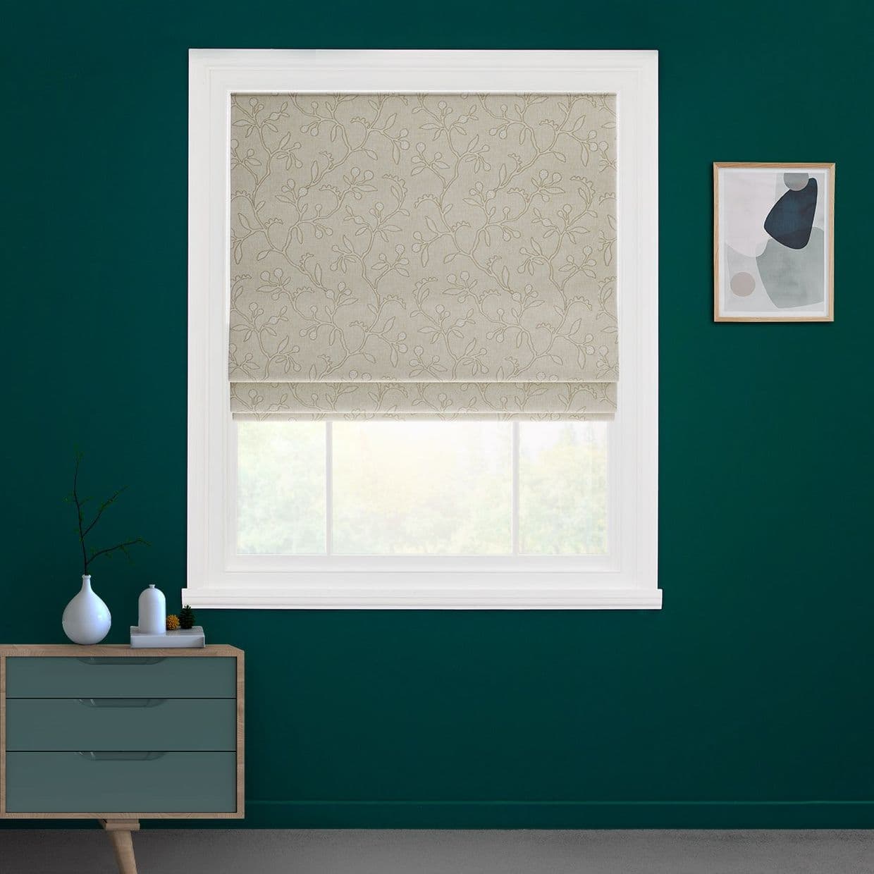Beige Roman blind with embroidered floral pattern, lowered over the window, filtering daylight; white trim on a deep teal wall, with a small dresser and a framed abstract print nearby.