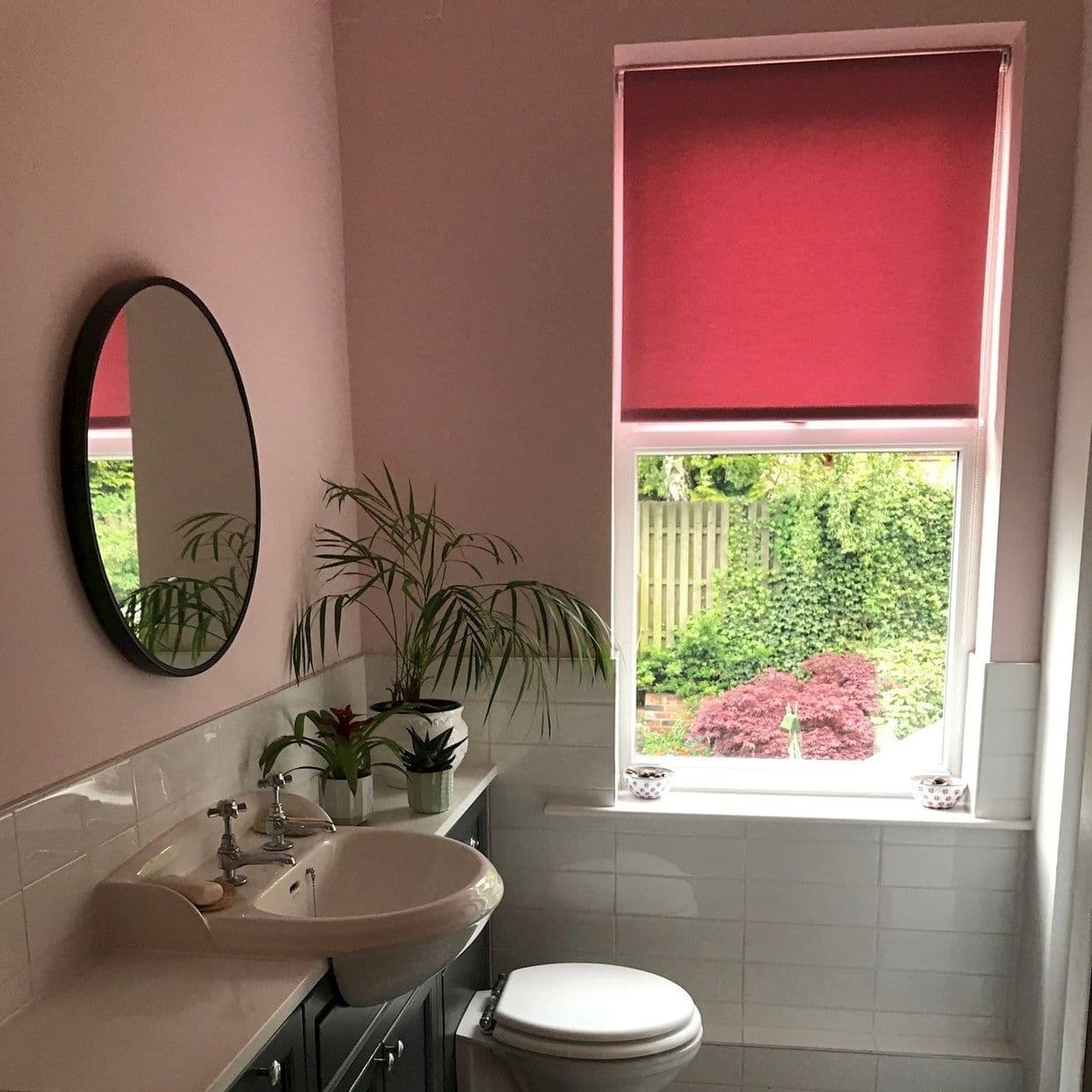 Red roller blind lowered over the top window, filtering warm light; bathroom interior with pink walls, round mirror, sink, potted plants, and a garden view through the lower pane.