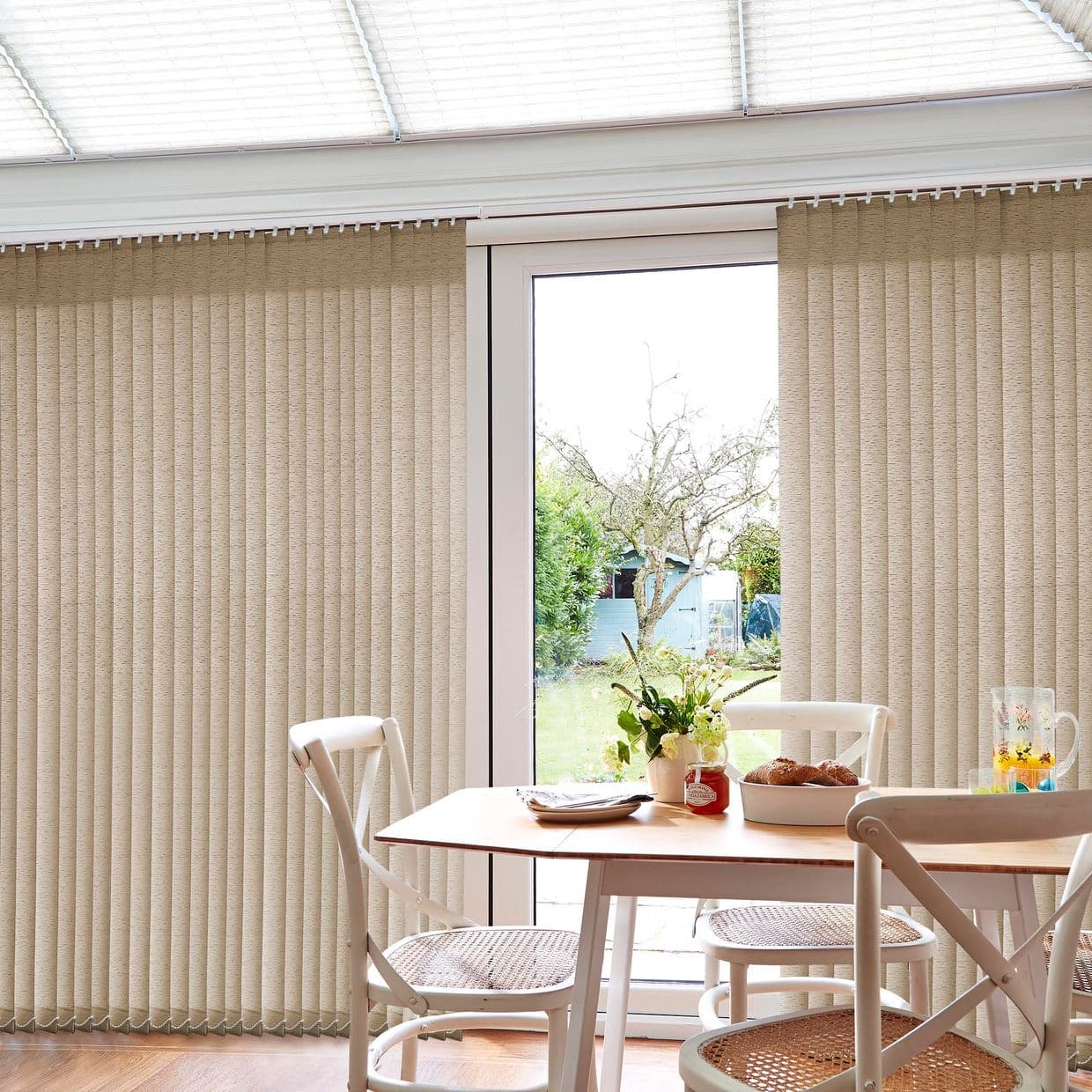 Beige vertical blinds (floor-to-ceiling) mostly closed with central gap, filtering daylight and revealing a glass patio door and garden beyond. Context: bright dining nook with wooden table, cane chairs, flowers.