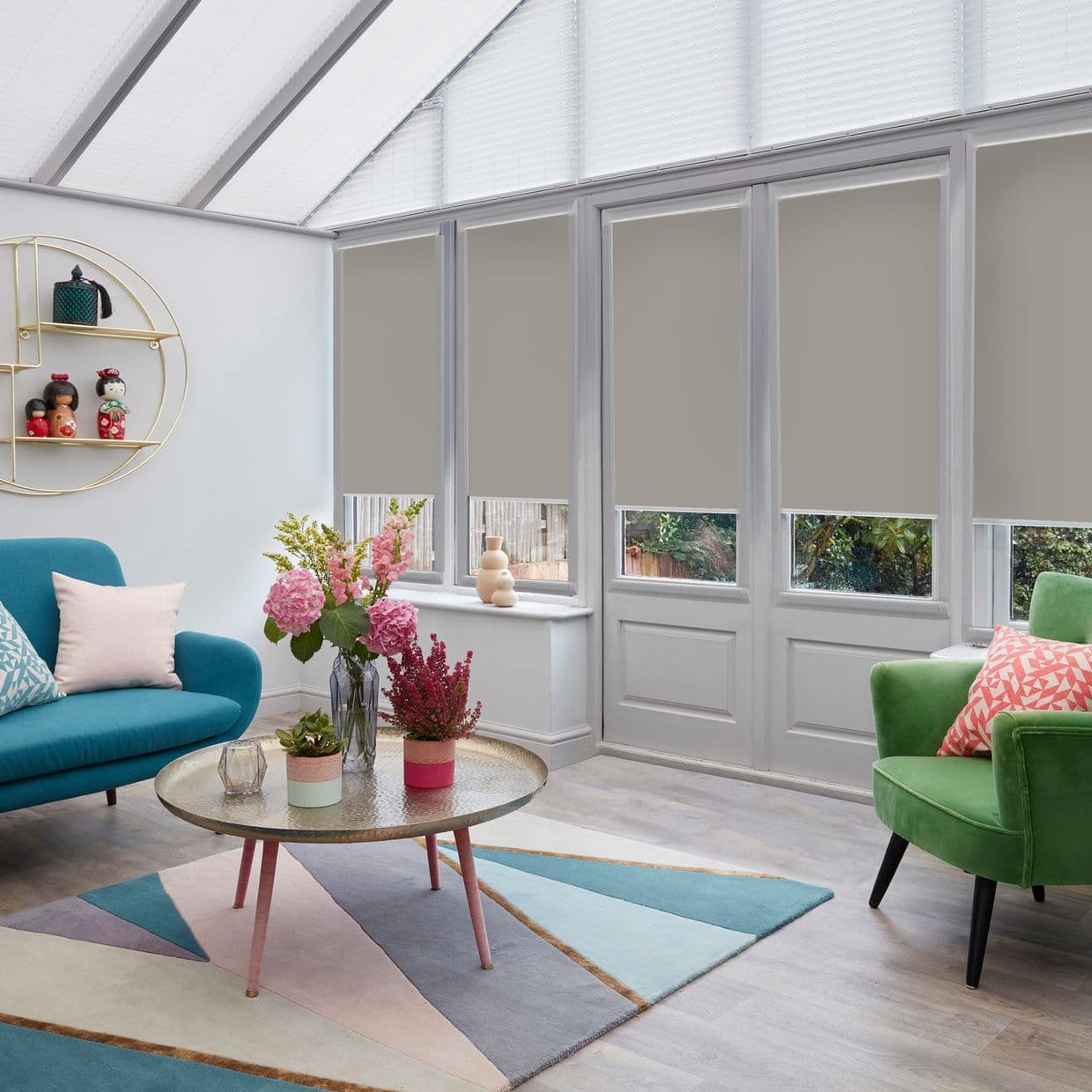 Grey roller blinds, lowered over tall glass doors and windows leaving clear bottom panels, filtering light and providing privacy in a bright conservatory-style living room with pastel furniture.