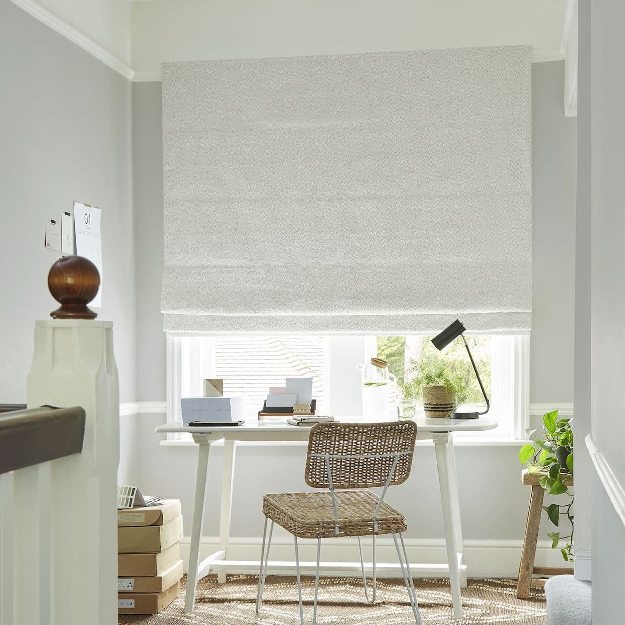 White textured Roman shade lowered over a window, blocking most light and letting soft daylight from the bottom; small home office desk with wicker chair, lamp and potted plants. 01