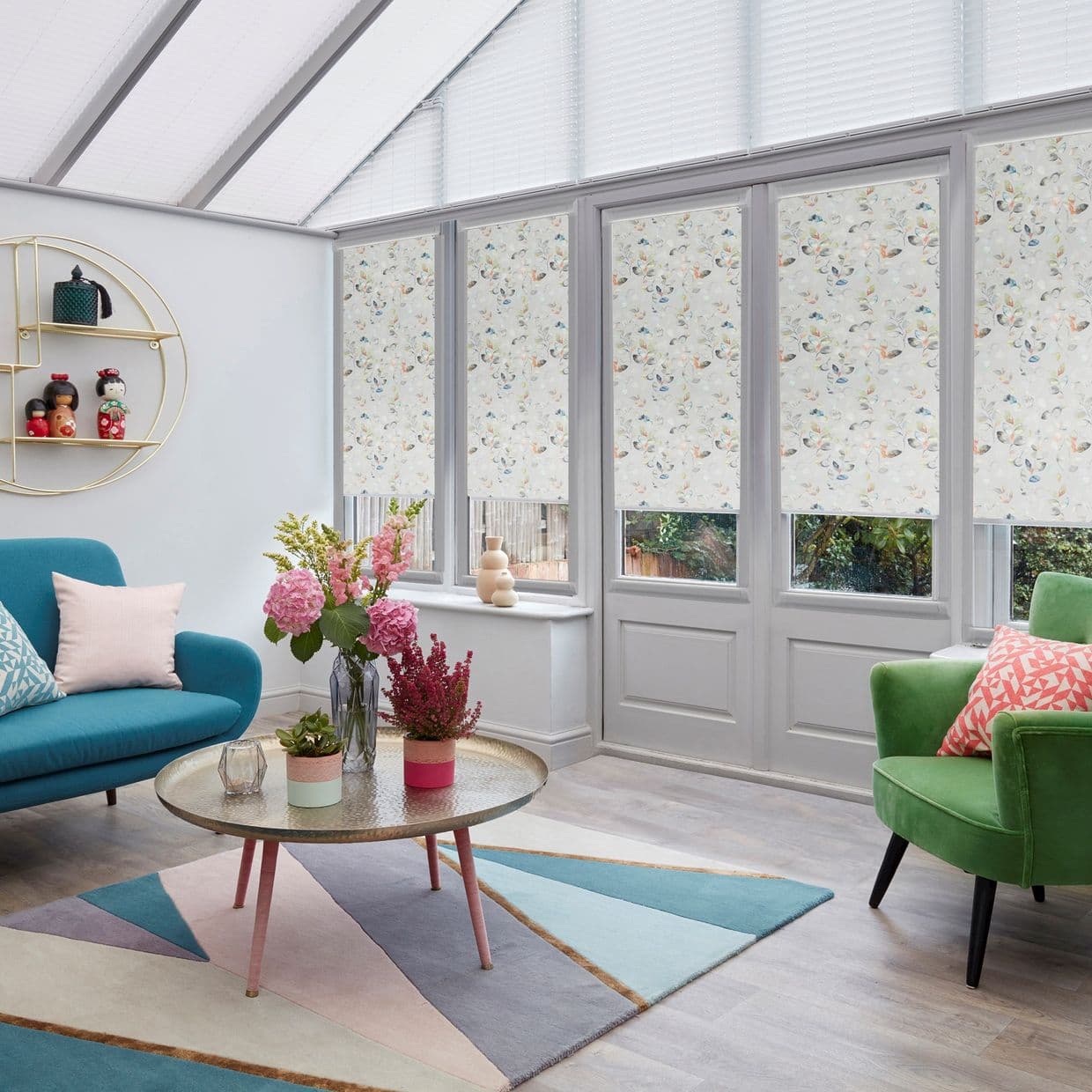 Floral-patterned roller blinds, lowered over tall French doors and windows with narrow clear lower panes, gently filtering daylight; in a bright conservatory living room with sofas, rug and flowers.