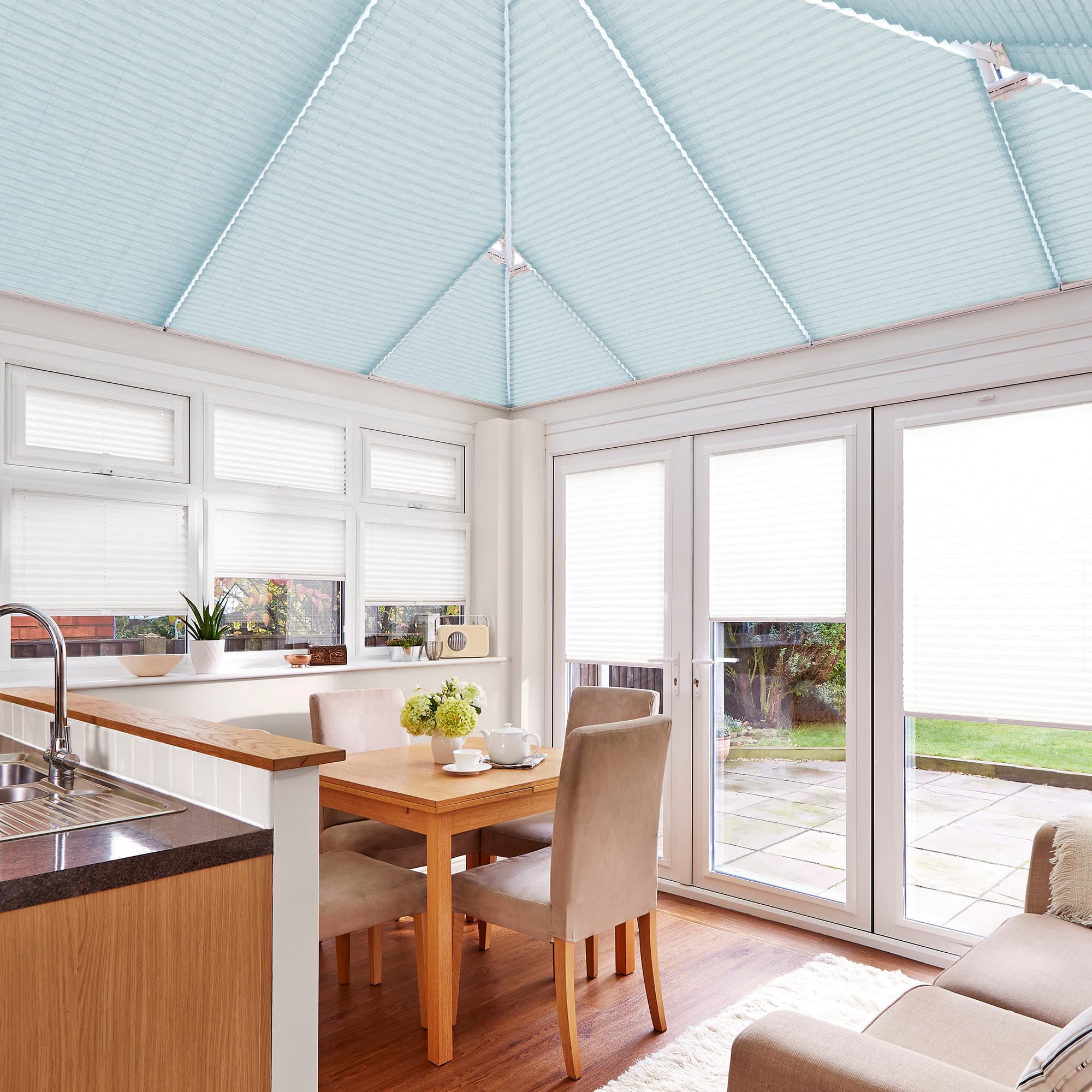 Pleated pale-blue roof blinds cover the conservatory ceiling, closed and diffusing light; white pleated window and door blinds partly lowered; bright dining-kitchen with wooden table, chairs and patio view.