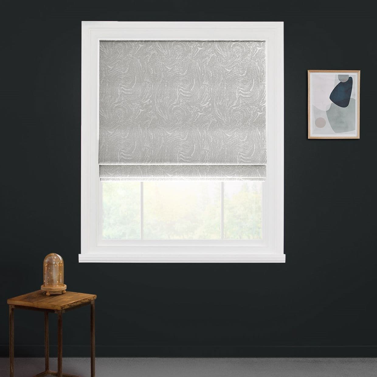 Textured gray Roman blind, lowered to cover most of a white window, gently filtering daylight through a gap; against a dark charcoal wall with a side table and abstract framed print.