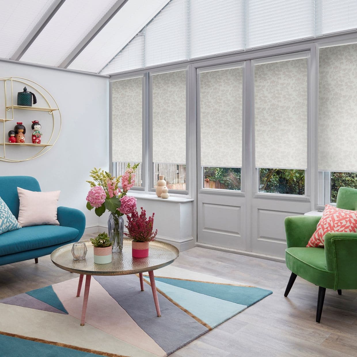 Roller blinds with pale floral pattern, mostly lowered over tall conservatory windows, filtering soft daylight. Context: bright sunroom living area with teal sofa, green armchair, geometric rug and coffee table.