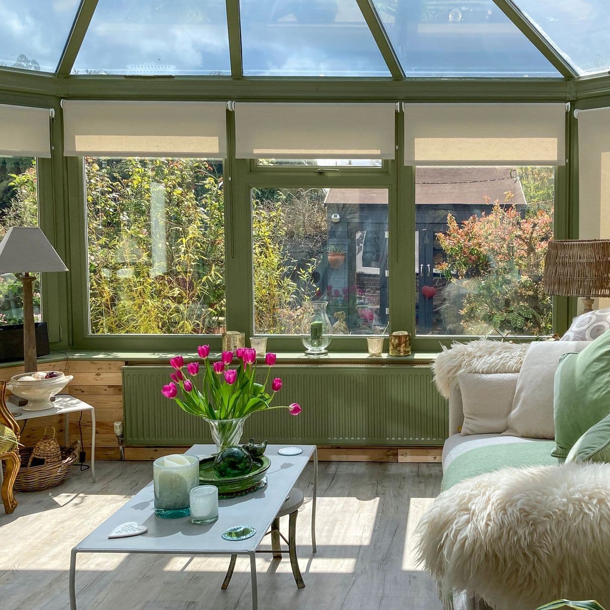 Cream roller blinds, partially lowered across green-framed conservatory windows, filtering sunlight; cozy sunlit sitting area with sofa, coffee table holding bright pink tulips and a leafy garden beyond.