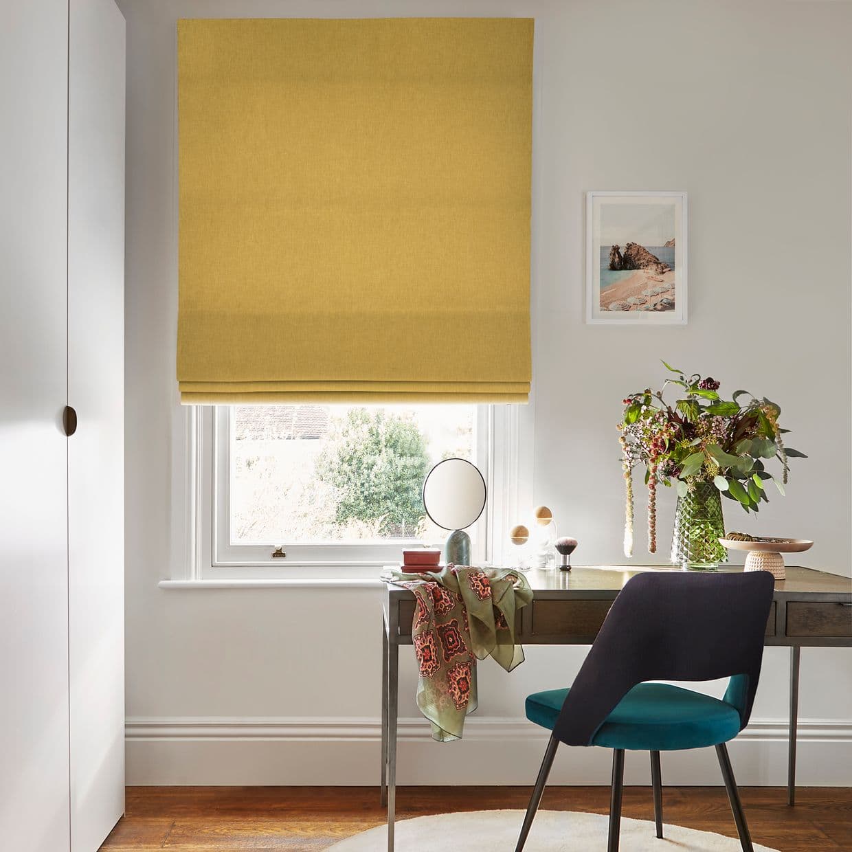 Yellow Roman blind covering most of a window, fully lowered and folded at the bottom, filtering soft daylight into a tidy home office with desk, chair, vase and framed print.