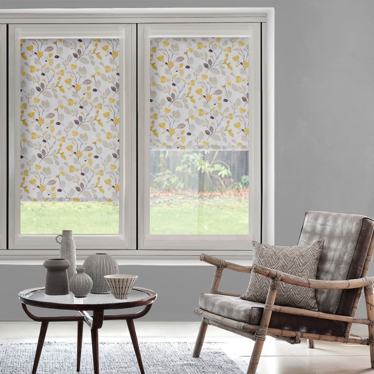 Floral roller blinds (white with yellow and beige leaf pattern) are partially lowered, filtering daylight; framed windows reveal a grassy backyard, inside a gray living room with wooden chair and ceramics.
