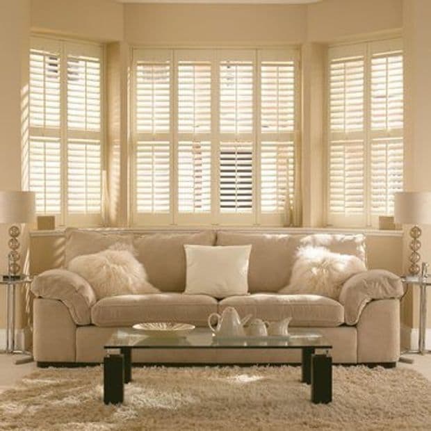 Cream-colored wooden plantation shutters covering a three-section bay window, slats partially angled to filter warm daylight; beige living room with plush sofa, fluffy cushions, glass coffee table and shag rug.