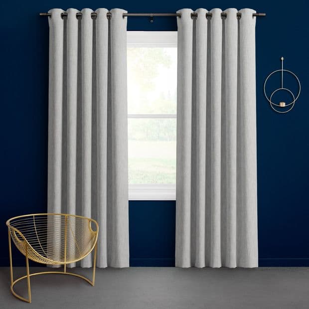 Object: Light gray grommet-top curtains hung in even pleats, parted to reveal a central white-framed window. Action: Framing and softly filtering bright daylight. Context: Deep navy wall with gold wire chair.