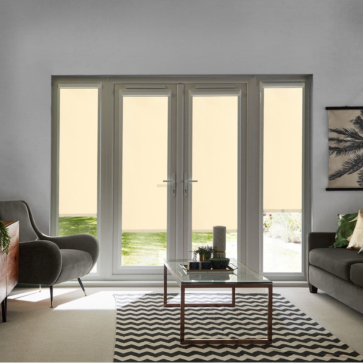 Beige roller blinds covering French doors and sidelights, lowered nearly fully to block view and filter daylight, leaving narrow bottom panels revealing lawn; living room with glass coffee table, sofas.