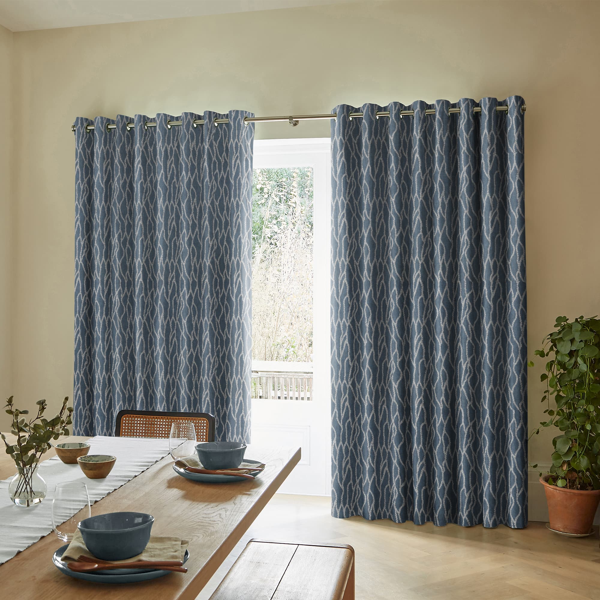 Blue patterned grommet-top curtains, hung on a metal rod, are partially drawn and filtering daylight through a central gap; dining room with wooden table settings, neutral walls, and a potted plant.