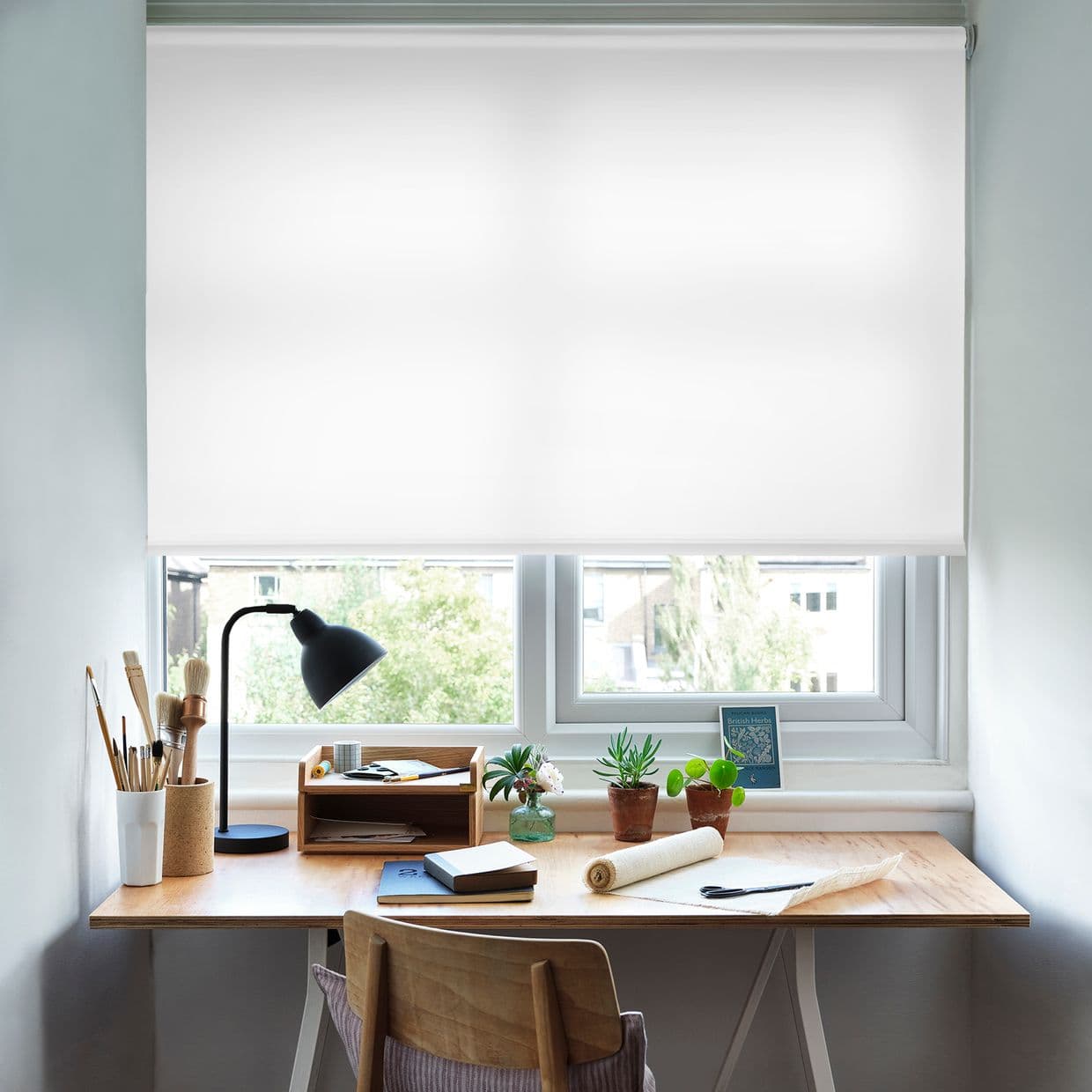 A white roller blind, fully lowered and translucent, filtering bright daylight; a wooden desk beneath with lamp, brushes, notebooks and potted succulents; compact home workspace facing trees and houses outside.