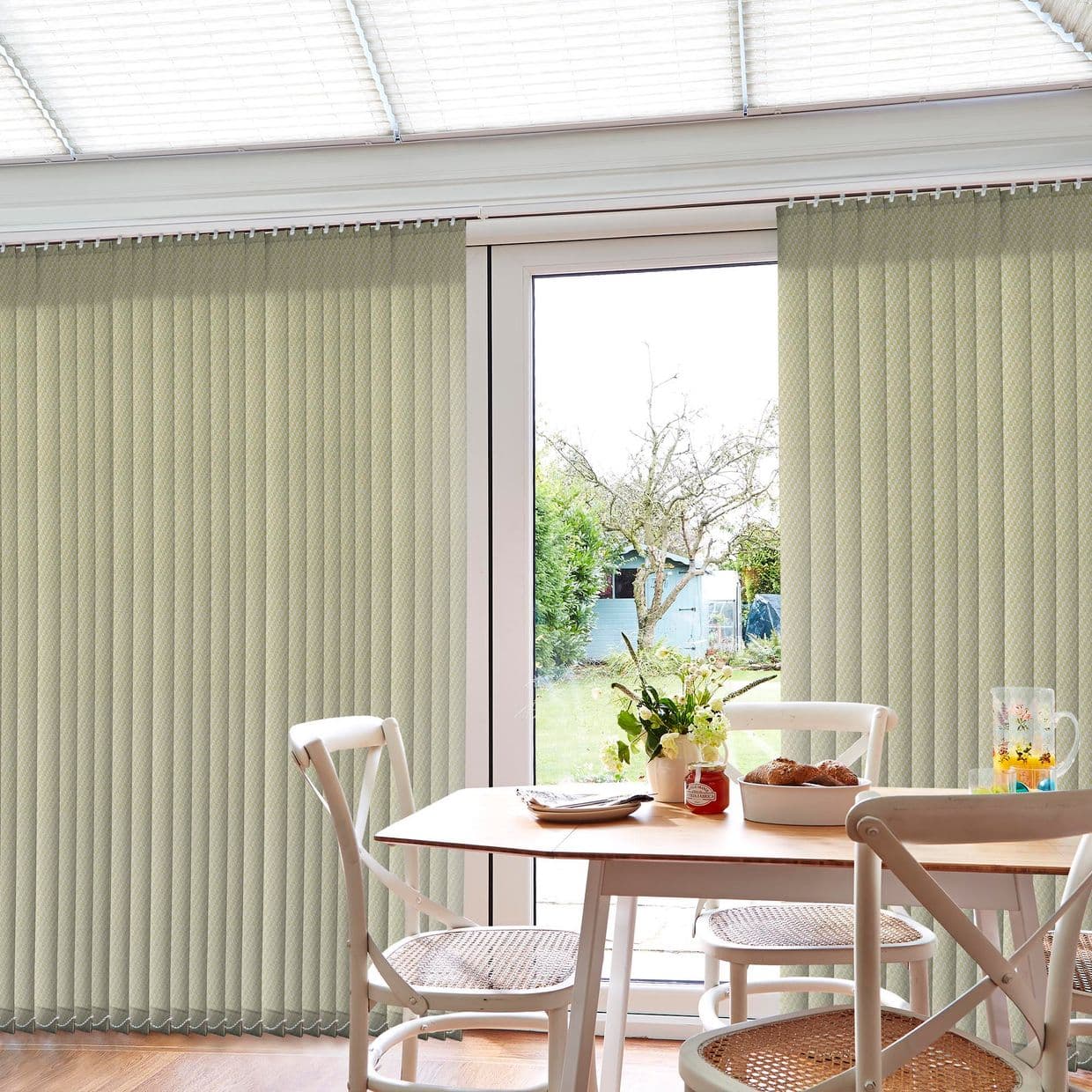Beige vertical blinds across a sliding glass door, mostly closed with a central garden gap; filtering daylight into a dining nook with wooden table, white chairs, vase.