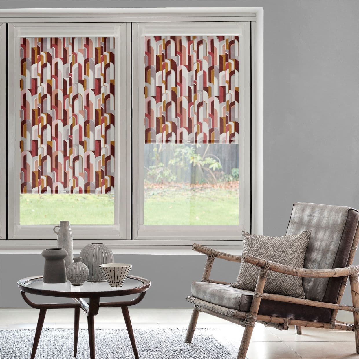 Patterned roller blinds on two adjacent windows, half-lowered, multicolored arch print; filtering light and obscuring upper view; in a neutral living room with rattan leather armchair, round table, garden beyond.