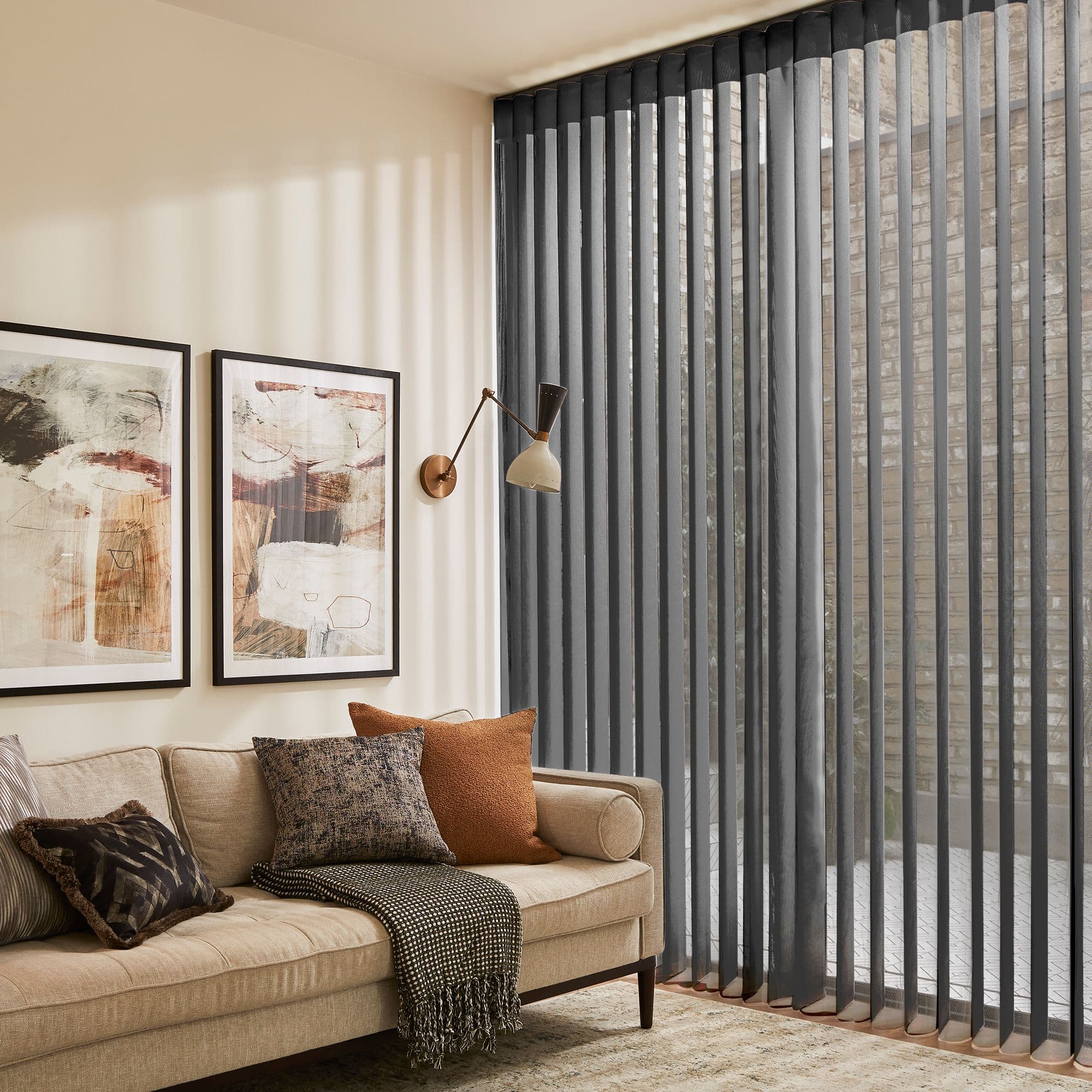 Dark grey vertical blinds, floor-to-ceiling sheer slats along a corner windowâ€”filtering daylight and casting striped shadowsâ€” in a warmly lit living room with beige sofa, framed art, and wall lamp.