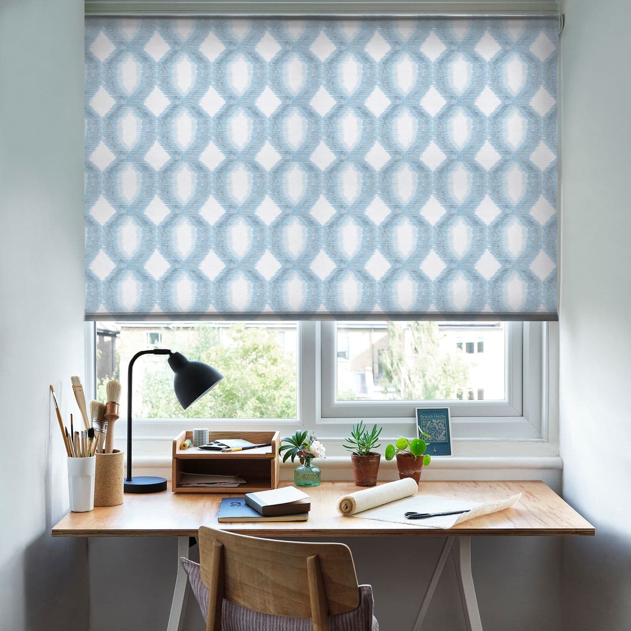 Patterned roller blind (blue-white interlocking oval motif) pulled down, filtering daylight over a small home desk; wooden workspace with lamp, plants, art supplies, and a leafy outside view.
