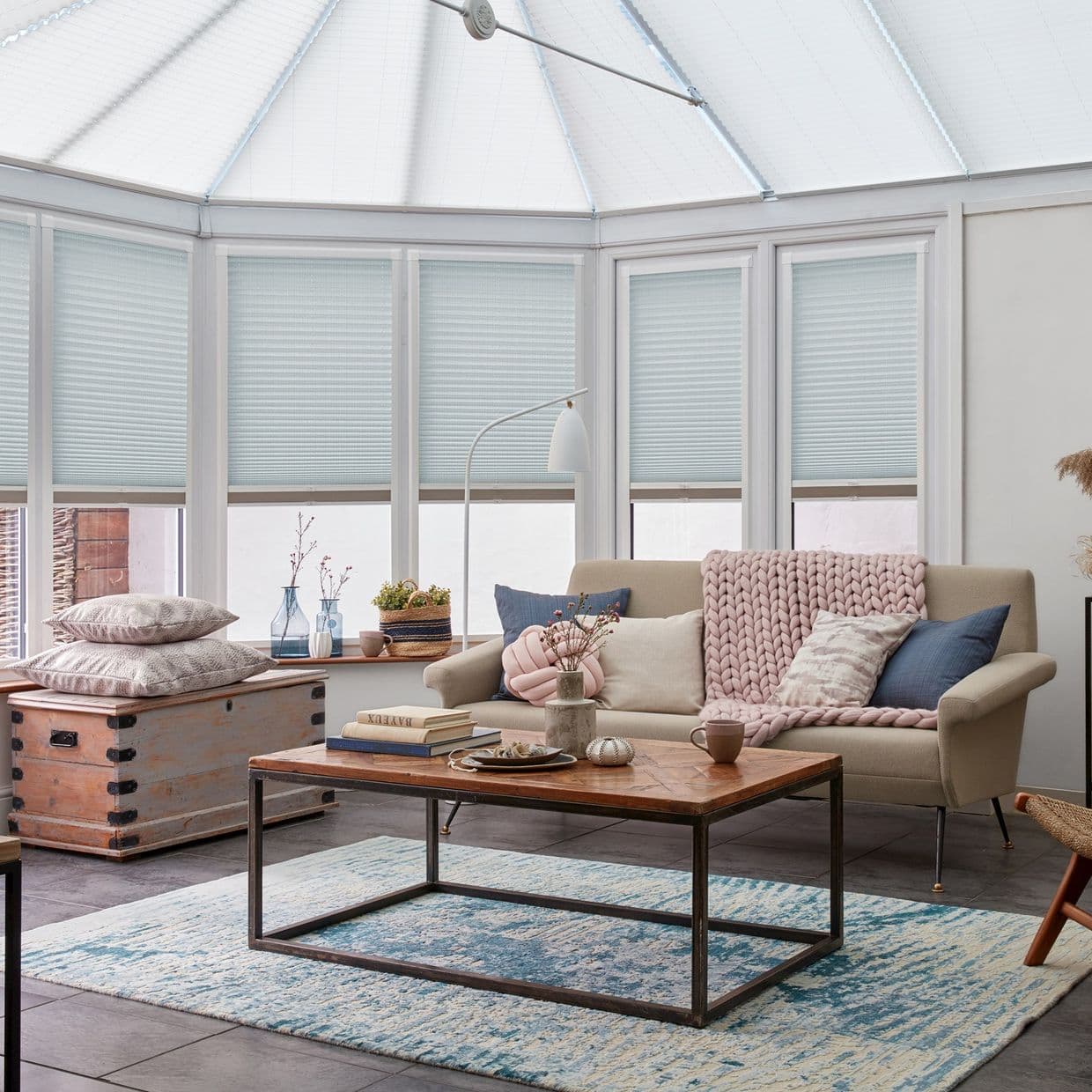 Pale blue cellular shades lowered to mid-height across conservatory windows, filtering soft daylight; beige sofa with chunky pink throw, wooden coffee table on a blue rug in a tiled sunroom.