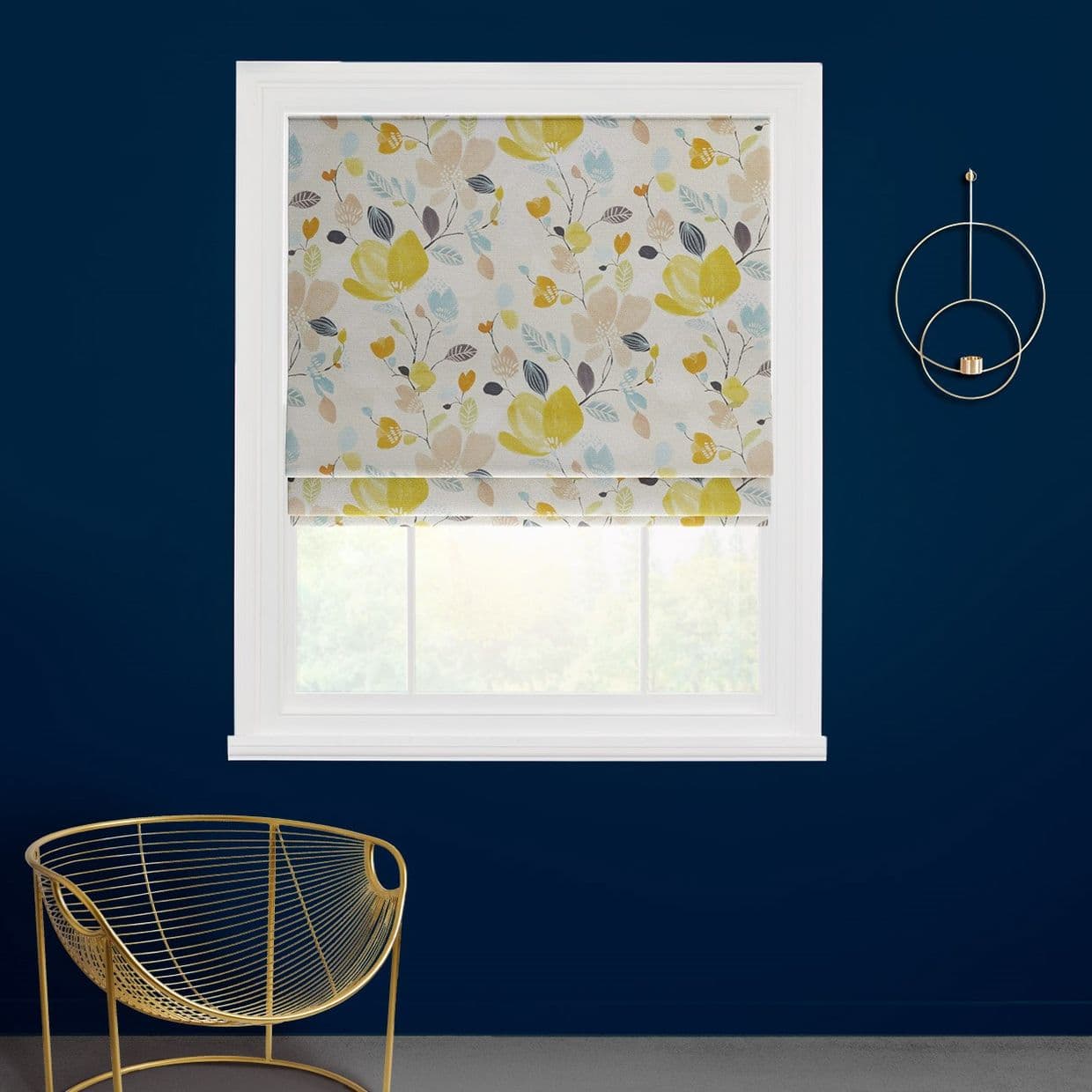 A floral Roman blind with yellow, blue and beige blossoms covers a window, filtering daylight, in a navy-walled room with white frame, gold wire chair and circular wall candleholder.