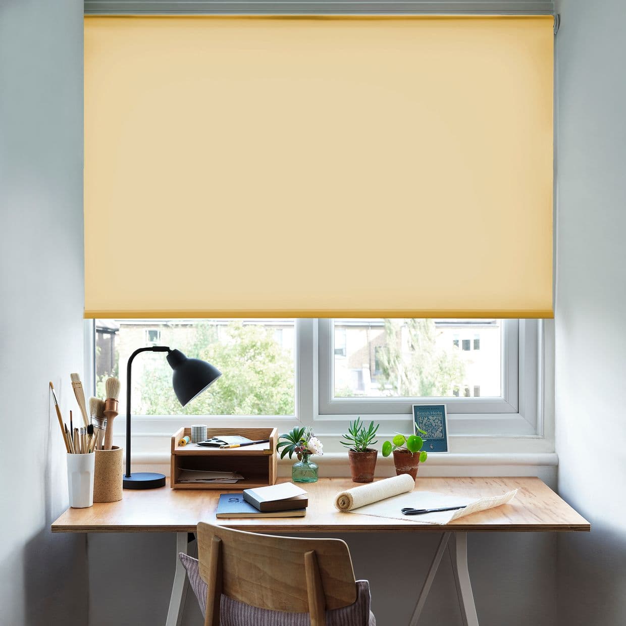 Yellow fabric roller blind lowered over a double window, filtering soft daylight; a wooden desk beneath holds a black desk lamp, notebooks, paintbrushes and potted succulents in a bright workspace.