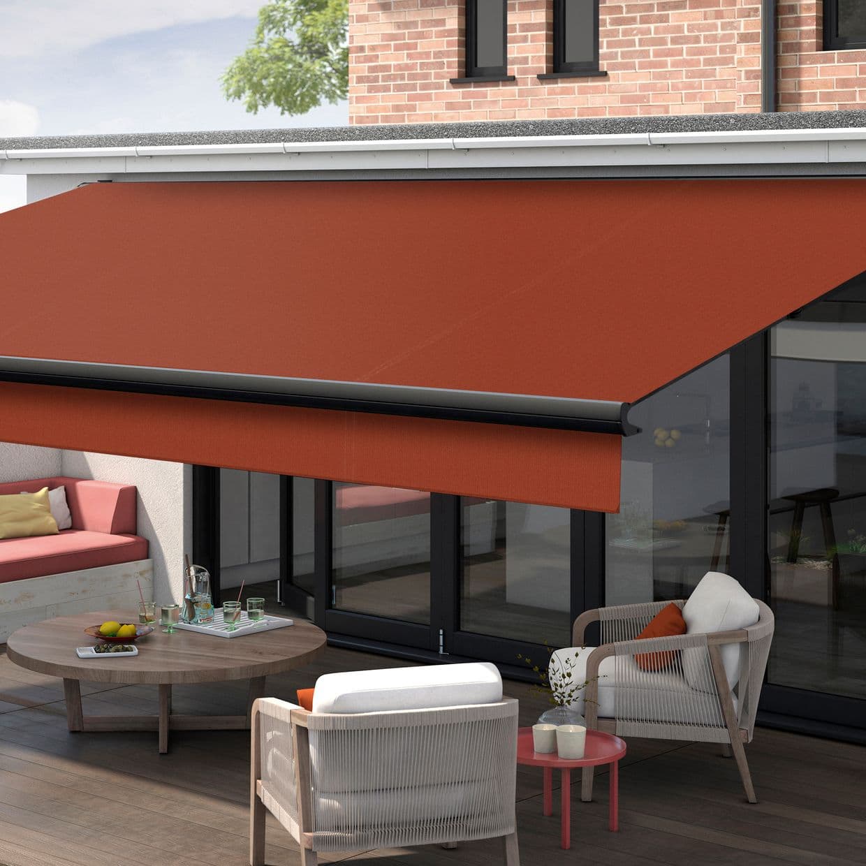 Red retractable awning with black casing, fully extended over glass folding doors, shading a wooden-deck patio with lounge chairs, round coffee table, and brick-backed exterior in daylight.