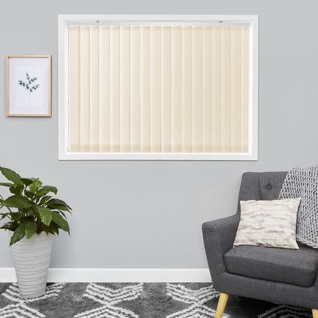 Beige vertical blinds covering a window, fully closed and filtering daylight; set in a gray living room above a patterned rug, beside a potted plant and a gray upholstered armchair.