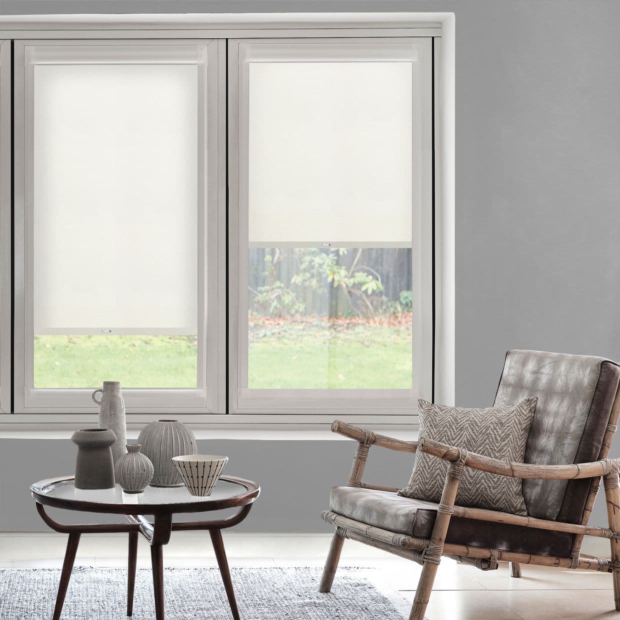 White roller blinds inside a double window; left fully lowered, right partially, filtering soft daylight. Gray living area with rattan leather armchair, patterned cushion, round coffee table with ceramics; lawn beyond.