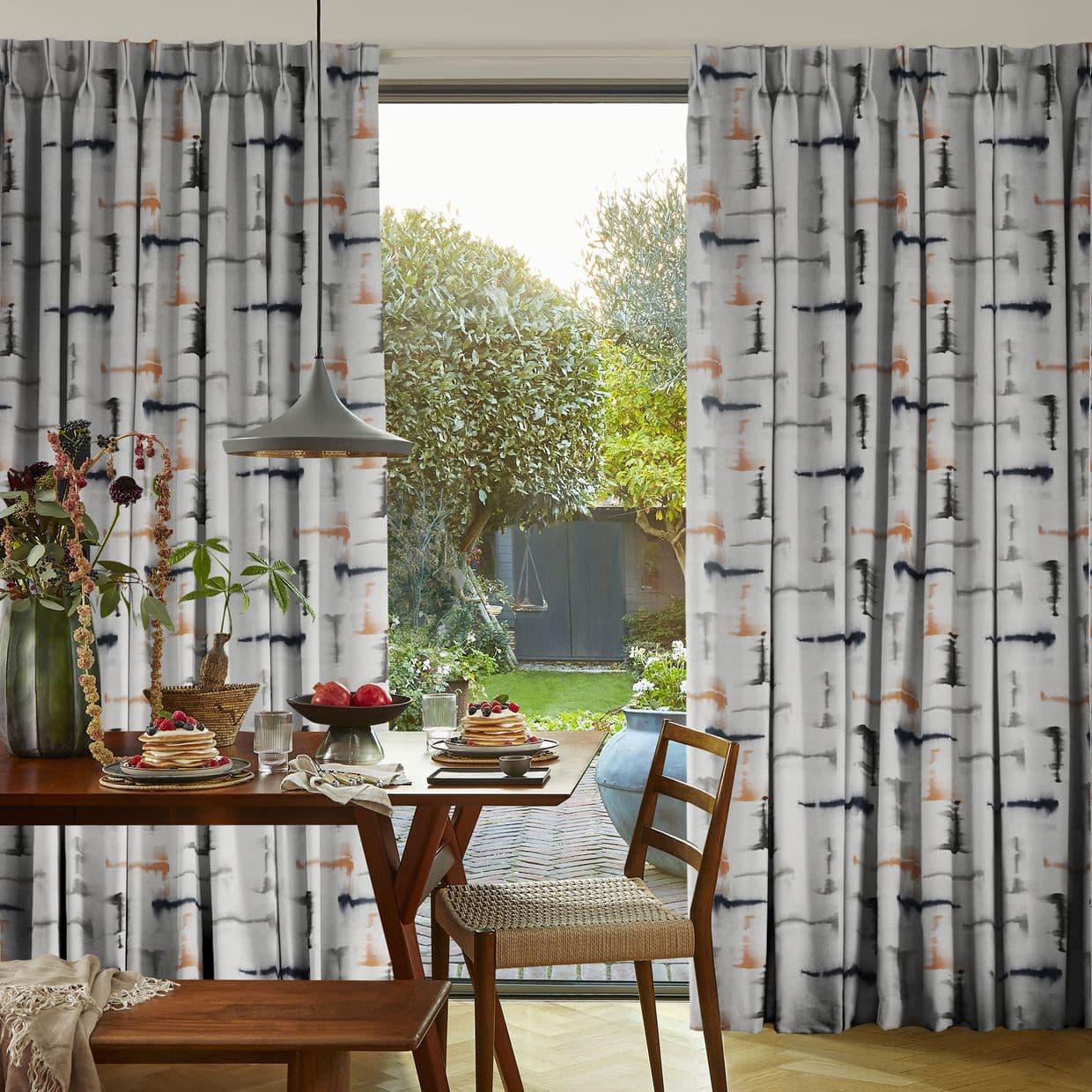 Pleated patterned curtains (grey with black and rust brushstrokes) pulled aside at a central sliding door, framing a sunlit backyard; foreground dining table set with pancakes, pendant lamp, wooden chairs.