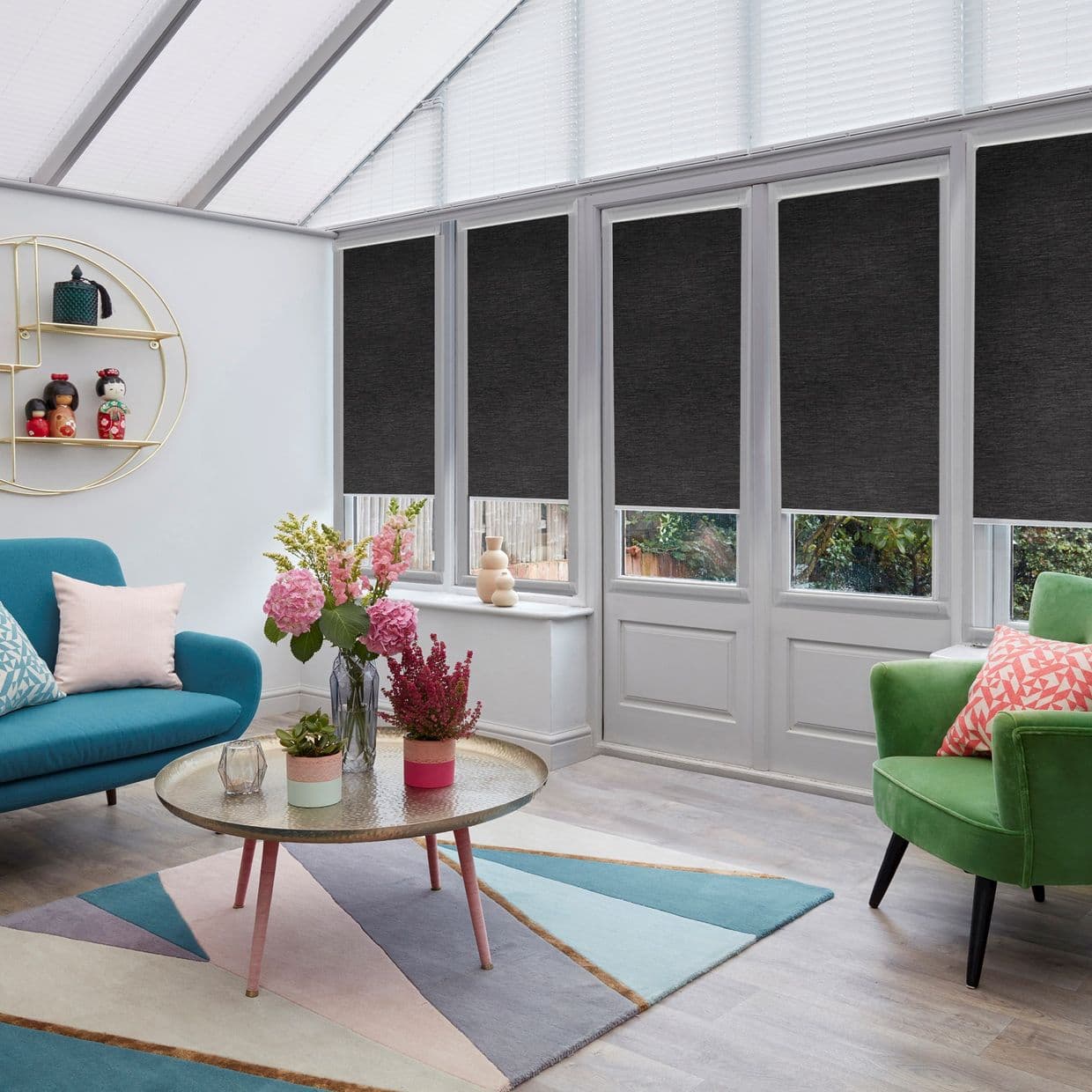 Dark grey roller blinds, lowered across tall conservatory windows and French doors, blocking upper light while leaving lower panes exposed; bright sunroom furnished with teal sofa, green armchair, coffee table.