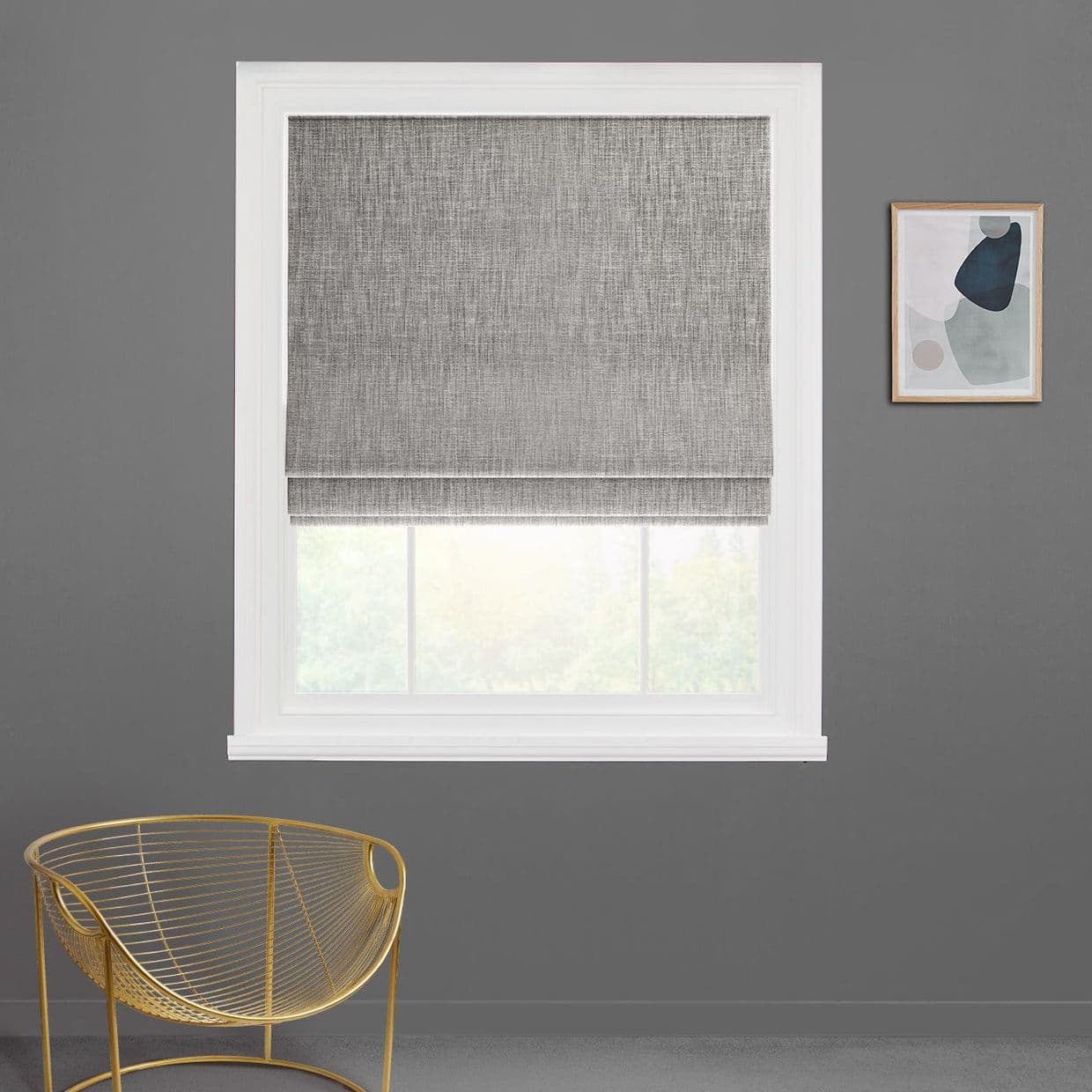 Gray Roman shade, mostly lowered with a small bottom gap filtering light; set in a white-framed window on a gray wall beside a gold wire chair and abstract print.